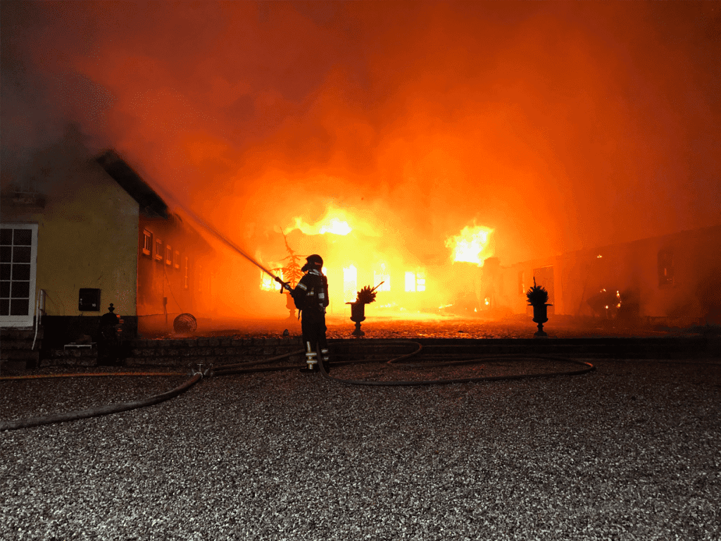 En brandmand i silhuet foran en stor bygningsbrand, mens han sprøjter vand mod flammerne med brandslange.